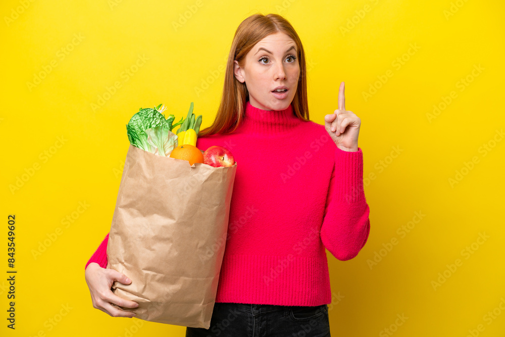Young redhead woman holding a grocery shopping bag isolated on yellow background thinking an idea pointing the finger up Young redhead woman holding a grocery shopping bag isolated on yellow background thinking an idea pointing the finger up