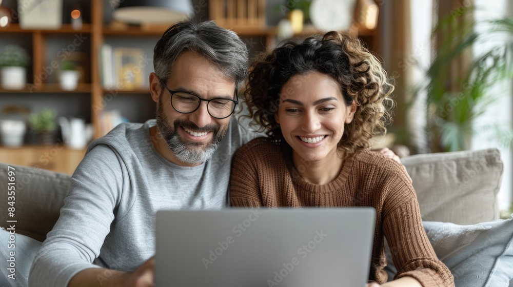 A couple sits on a couch in their living room, smiling while looking at a laptop together A couple sits on a couch in their living room, smiling while looking at a laptop together