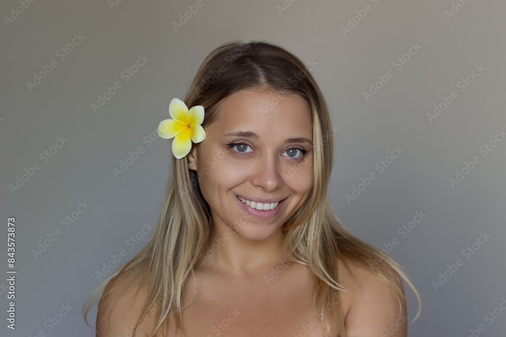 Portrait a young attractive smiling caucasian blonde woman with white and yellow frangipani flower in her hair on a dark gray background. Hair ornaments, decorations, beauty concept Portrait a young attractive smiling caucasian blonde woman with white and yellow frangipani flower in her hair on a dark gray background. Hair ornaments, decorations, beauty concept