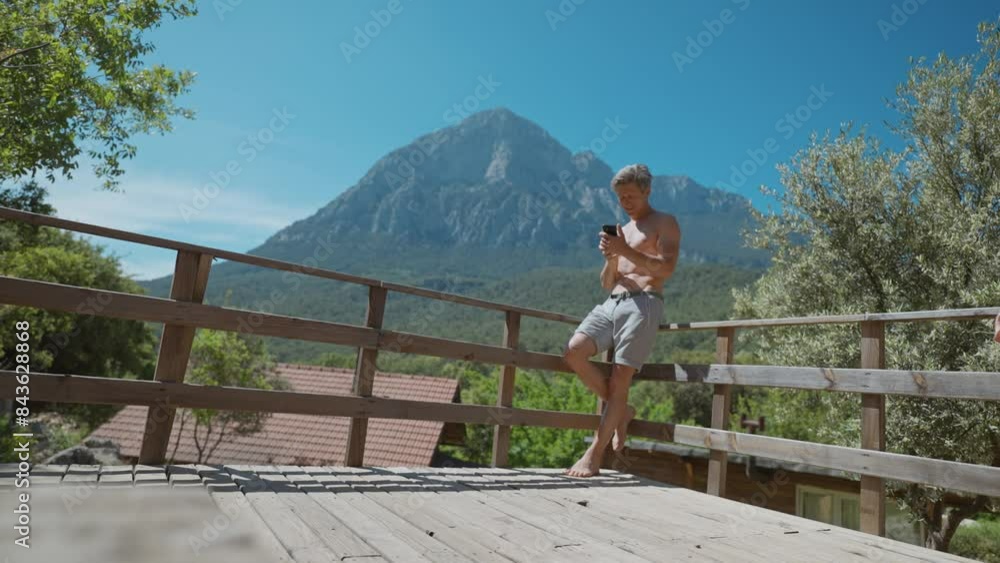 Adult shirtless man with fit athletic body enjoying warm sunny day on wooden veranda with majestic scenic mountains on background. smiling man using phone outdoors for taking photo and video. Adult shirtless man with fit athletic body enjoying warm sunny day on wooden veranda with majestic scenic mountains on background. smiling man using phone outdoors for taking photo and video.