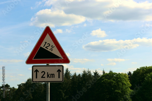 Verkehrsschild Achtung Gefälle, Verkehrszeichen Nr. 108, mit Hinweisschild "2 km" in der Natur vor blauem Himmel.