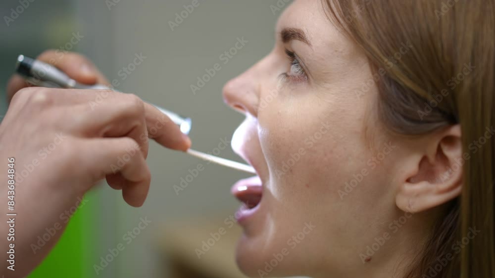 Close-up. A doctor asks a female patient to open mouth for a visual examination of the mucous membrane of the throat. The doctor uses a wooden spatula, to press the back of the tongue and a medical Close-up. A doctor asks a female patient to open mouth for a visual examination of the mucous membrane of the throat. The doctor uses a wooden spatula, to press the back of the tongue and a medical