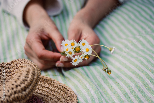 A bouquet of field white daisies plucked in a spring meadow. A young woman lying on a striped plaid with a bouquet of wildflowers. Straw female sunhat