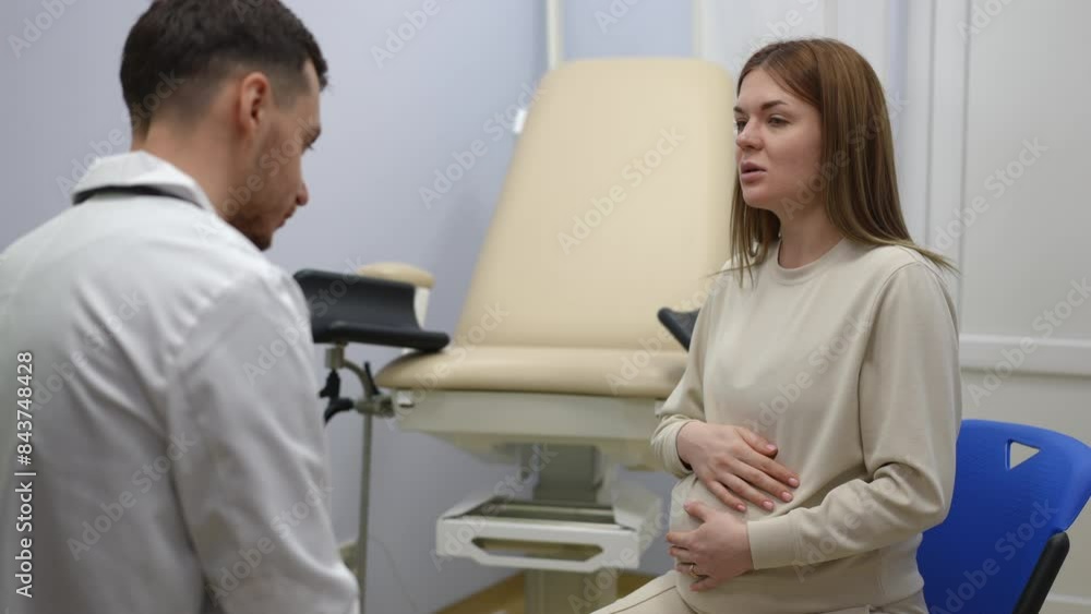 Slow motion. A pregnant woman, holding her belly with her hands, tells about her health problems to a male doctor. Doctor and patient sitting on chairs near the gynecological chair in the emergency Slow motion. A pregnant woman, holding her belly with her hands, tells about her health problems to a male doctor. Doctor and patient sitting on chairs near the gynecological chair in the emergency