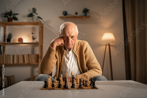 Serious senior man sitting in evening living room and playing chess in lonely. Solitude and retirement photo