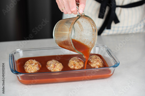 Female chef in an apron preparing meat or cutlets patties with rice, baking them in a dish with tomato sauce. Home cooking, traditional recipes, and culinary preparation.