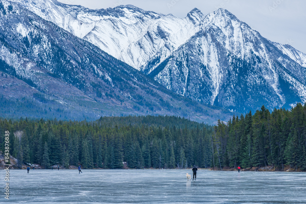 Tourists doing ice-skating in Johnson Lake frozen water surface in winter time. Snow-covered mountain in the background. Banff National Park, Canadian Rockies, Alberta, Canada. Tourists doing ice-skating in Johnson Lake frozen water surface in winter time. Snow-covered mountain in the background. Banff National Park, Canadian Rockies, Alberta, Canada.
