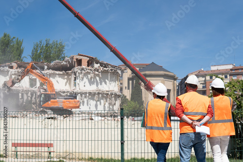 three construction engineers with blueprints and dressed in orange vests and hard hats watch as a bulldozer destroys a residential building in the city next to a park. copy space.