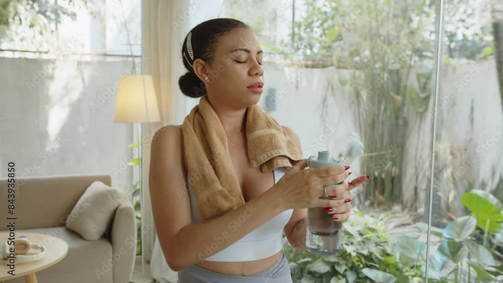 Medium shot of young Latin woman drinking water from plastic bottle and wiping sweat with towel after home training, copy space