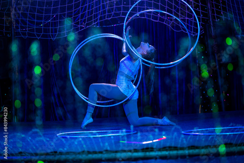 Young slim attractive circus gymnast woman performing live show with hula hoops on red arena with blue backlight, audience is excited