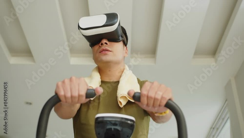 Low angle of young man using augmented reality headset when sitting on exercise bike, working out at home