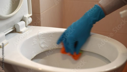 Close-up hands of unrecognizable househusband in gloves cleaning toilet bowl in bathroom. Housewife male doing cleaning and washing toilet bowl with sponge. Concept of domestic work, housekeeping.