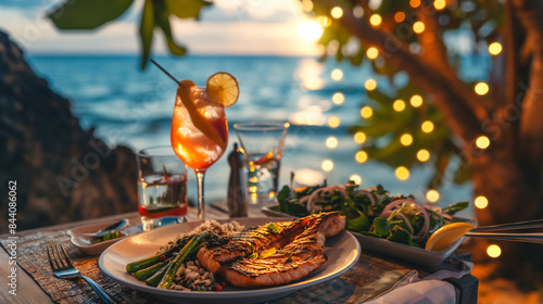 A picturesque table set by the beach with a gourmet meal of grilled swordfish, asparagus, and wild rice, accompanied by a tropical cocktail, under a canopy of string lights photo