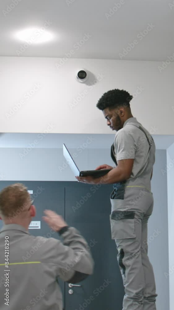 Two multi ethnic installers in uniform sets up security cameras in office corridor using laptop. Upset African American worker looks at camera. Concept of surveillance systems and privacy. Quick zoom Two multi ethnic installers in uniform sets up security cameras in office corridor using laptop. Upset African American worker looks at camera. Concept of surveillance systems and privacy. Quick zoom