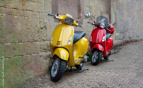 Two vintage scooter yellow, red color motorbike parked on paved pavement, stonewall background.