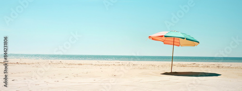 Beach umbrella with pastel stripes on a sandy beach against a pale blue sky background. Summer concept banner with copy space