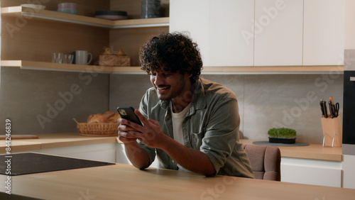 Food order carefree happy Indian man browsing smartphone laugh Arabian male smiling scrolling social media network app looking at mobile phone laughing smile enjoy funny video online at home kitchen