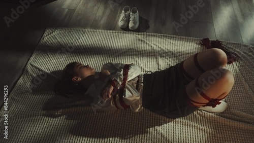 Woman lying on rug practicing art of Shibari with red ropes. Theme of restraint and vulnerability. White sneakers placed aside. Concept of artistic expression and tradition
