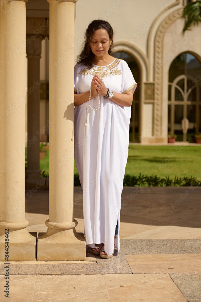 Fototapeta premium Woman in white dress praying outdoors in front of architectural pillars on a sunny day