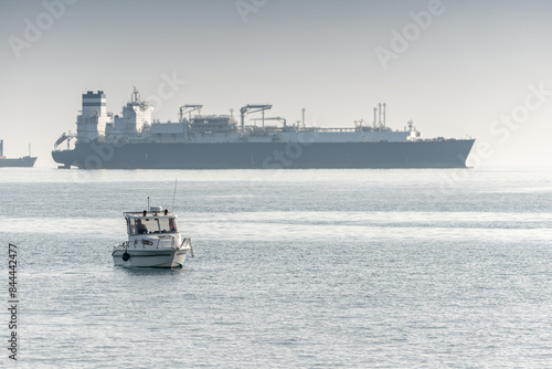 Small boat sailing near a large container ship on a hazy day at Akrotiri Bay, Cyprus