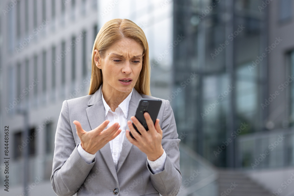 A professional woman looks upset while reading bad news on her phone. She stands outside a modern commercial building, wearing a grey suit. A professional woman looks upset while reading bad news on her phone. She stands outside a modern commercial building, wearing a grey suit.