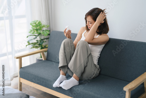 Young Woman Sitting on Sofa Holding Pill Bottle, Contemplating Medication, in Modern Minimalist Living Room with Natural Light and Green Plant