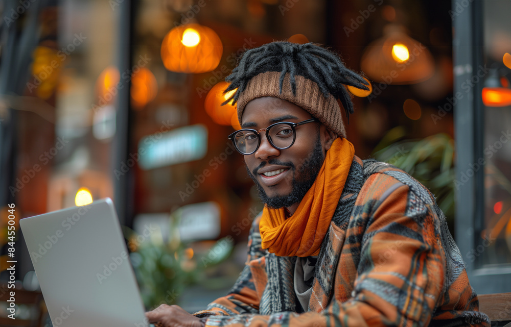 A young man wearing glasses is smiling and working on his laptop in a comfortable cafe setting A young man wearing glasses is smiling and working on his laptop in a comfortable cafe setting