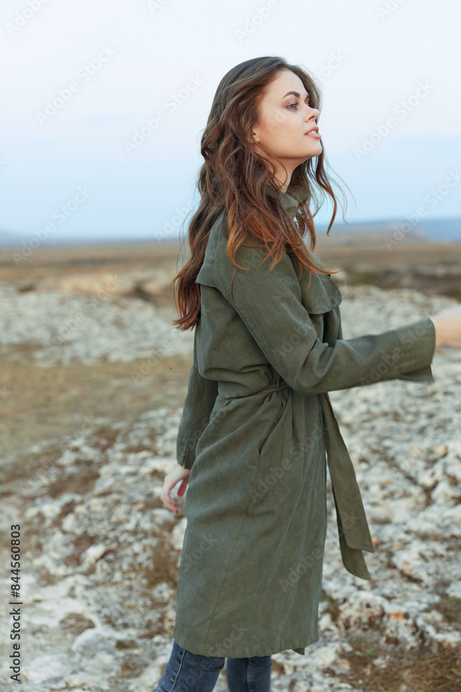 Woman in olive green trench coat standing on rocky hill overlooking ocean on sunny day Woman in olive green trench coat standing on rocky hill overlooking ocean on sunny day