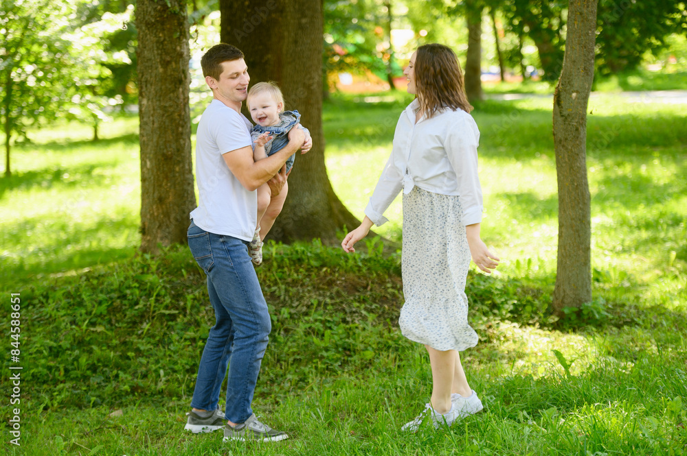 Fototapeta premium Joyful family plays with their baby in park on sunny day. Father holds girl in his arms and spins with her. Family weekend concept, happy moments