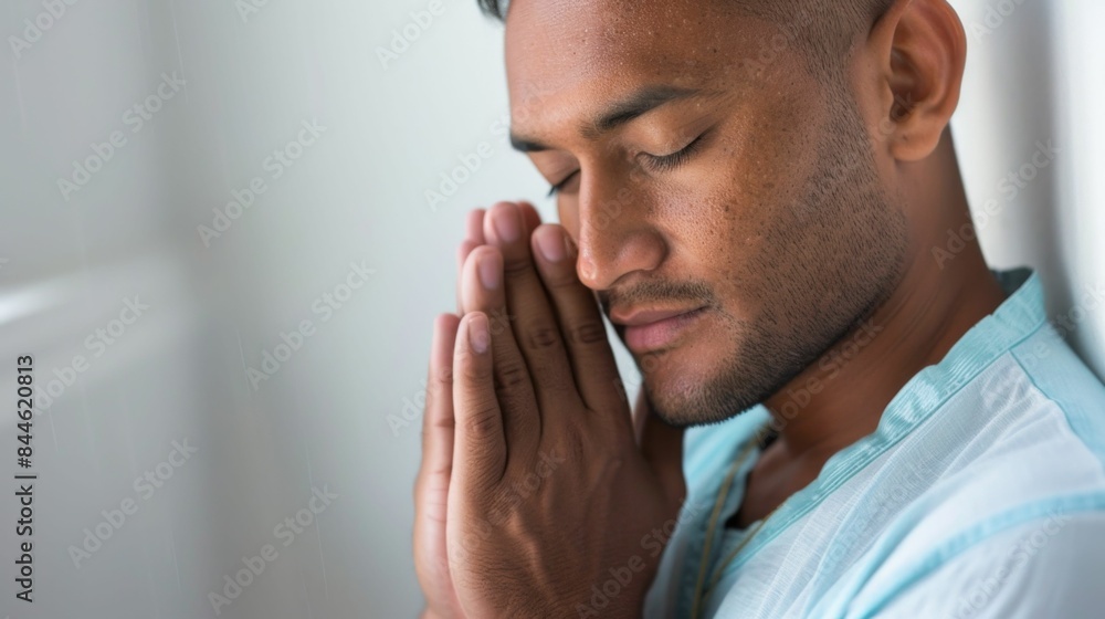 Fototapeta premium Man in blue shirt with eyes closed hands clasped in prayer standing against white wall.