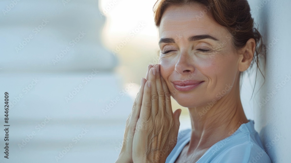 A woman with closed eyes smiling gently with her hands clasped together in front of her face against a blurred background. A woman with closed eyes smiling gently with her hands clasped together in front of her face against a blurred background.