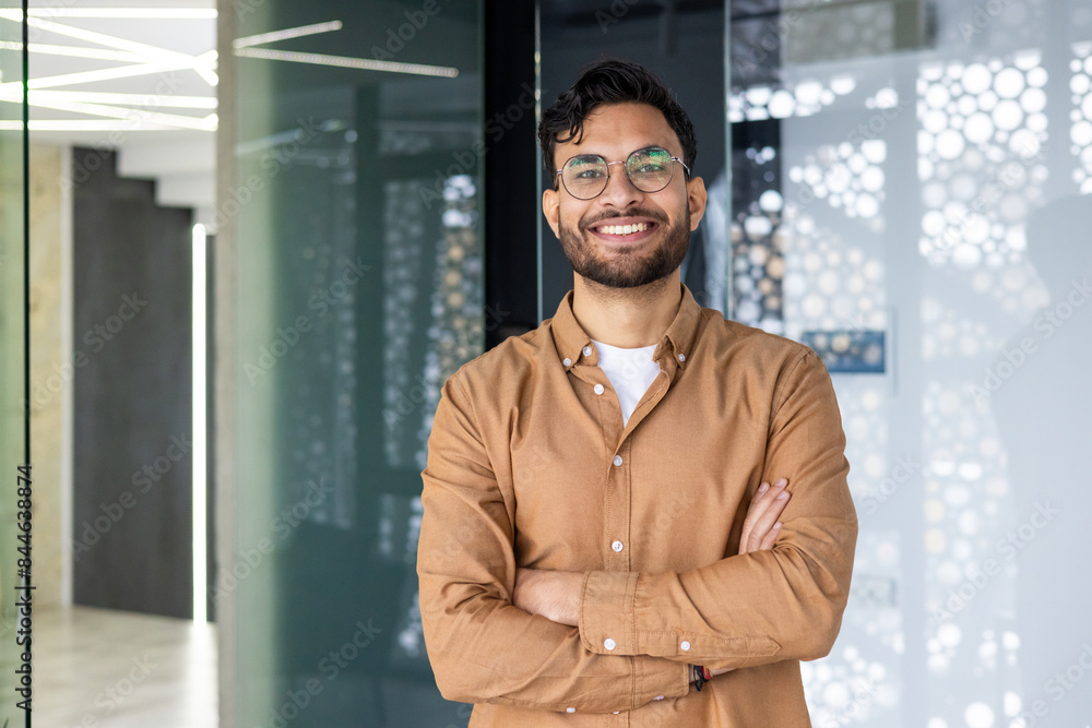 Naklejka premium Portrait of a young Indian man wearing glasses and a brown shirt standing in a modern office, crossing his arms over his chest and looking confidently at the camera