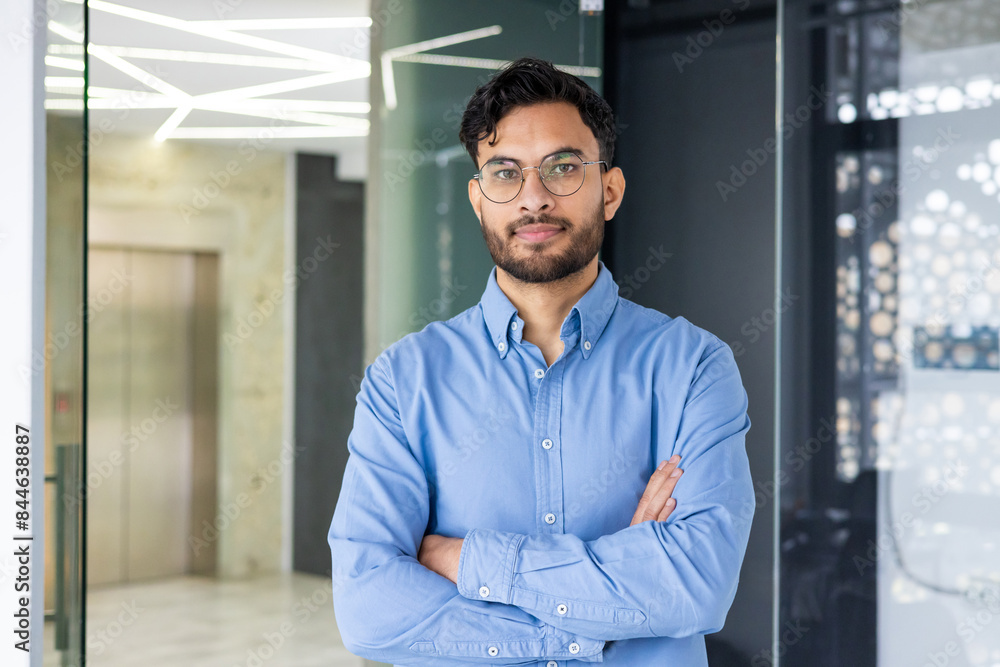 Fototapeta premium A serious Indian man in glasses and a blue shirt standing with crossed arms in a modern office and looking confidently into the camera