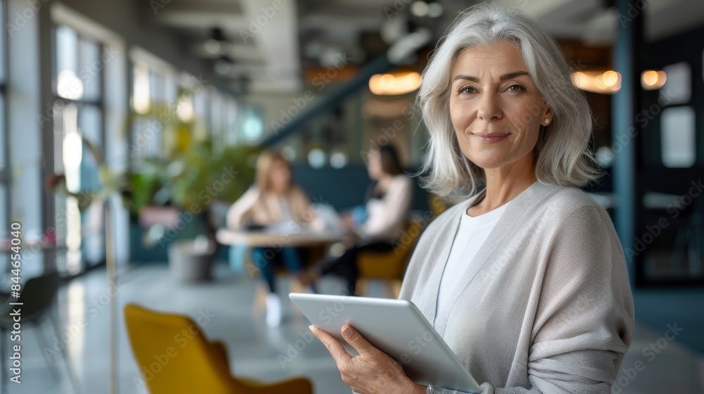 Business woman smiling at the camera while holding a tablet pc. Mature business woman standing in an office with her colleag Business woman smiling at the camera while holding a tablet pc. Mature business woman standing in an office with her colleag