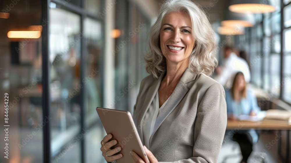 Business woman smiling at the camera while holding a tablet pc. Mature business woman standing in an office with her colleag Business woman smiling at the camera while holding a tablet pc. Mature business woman standing in an office with her colleag