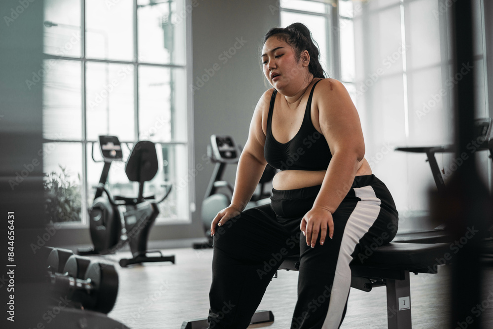Fototapeta premium Plus size Asian woman sitting on a bench in a gym, resting after an intense workout, reflecting dedication and physical exertion.