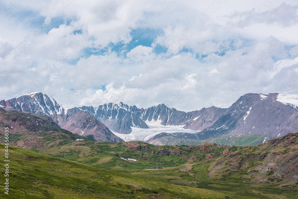 Fototapeta premium Scenic alpine landscape with green hills and rocks with view to big glacier and large snow-capped mountain range far away under clouds in blue sky in changeable weather. Awesome high snowy mountains.