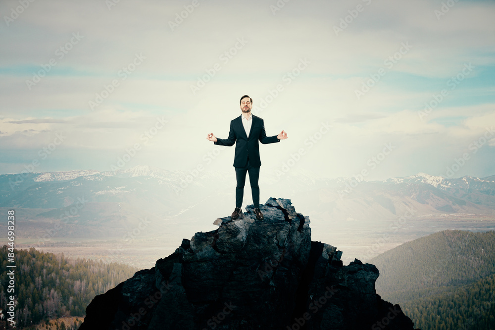 Attractive young man standing on mountain top and meditating. Bright sky background with mock up place. Success, peace and zen concept. Attractive young man standing on mountain top and meditating. Bright sky background with mock up place. Success, peace and zen concept.