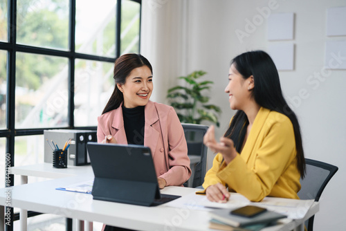 Two Professional Women Collaborating in Modern Office Setting with Laptops and Documents, Engaging in Productive Conversation, Business Meeting, Teamwork, and Corporate Environment © PaeGAG