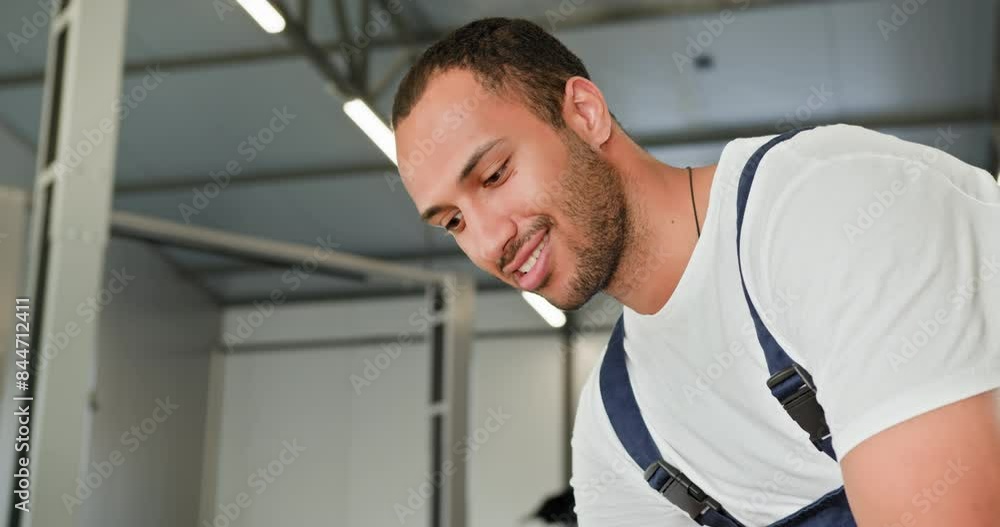 Auto Mechanic Working On A Repair In Workshop. A cheerful auto mechanic in a workshop, engaged in repairing a vehicle. He is wearing a white shirt and dark overalls, clearly focused on his work. Auto Mechanic Working On A Repair In Workshop. A cheerful auto mechanic in a workshop, engaged in repairing a vehicle. He is wearing a white shirt and dark overalls, clearly focused on his work.