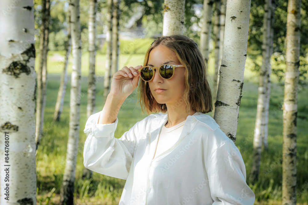 A stylish woman in a white shirt adjusts her sunglasses while standing in a birch tree forest. The sunlight and natural background reflect off her glasses, creating a cool and fashionable look. A stylish woman in a white shirt adjusts her sunglasses while standing in a birch tree forest. The sunlight and natural background reflect off her glasses, creating a cool and fashionable look.
