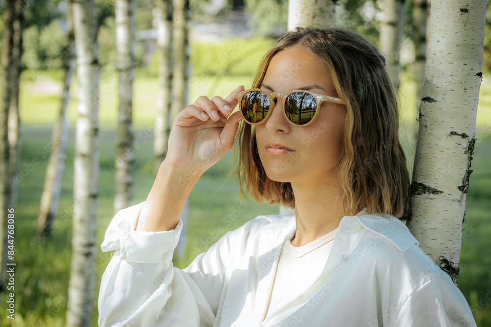 A stylish woman in a white shirt adjusts her sunglasses while standing in a birch tree forest. The sunlight and natural background reflect off her glasses, creating a cool and fashionable look. A stylish woman in a white shirt adjusts her sunglasses while standing in a birch tree forest. The sunlight and natural background reflect off her glasses, creating a cool and fashionable look.