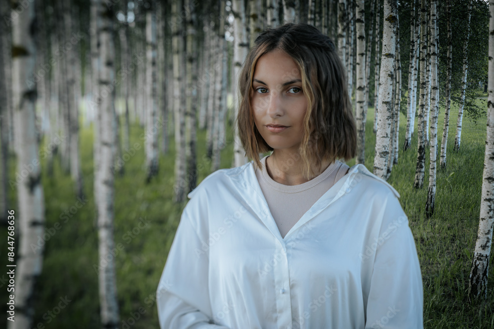 A woman with short hair and a calm expression stands in a dense birch tree forest, wearing a white shirt. The blurred background emphasizes the serene natural environment. A woman with short hair and a calm expression stands in a dense birch tree forest, wearing a white shirt. The blurred background emphasizes the serene natural environment.