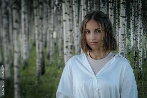 A woman with short hair and a calm expression stands in a dense birch tree forest, wearing a white shirt. The blurred background emphasizes the serene natural environment. © Emvats