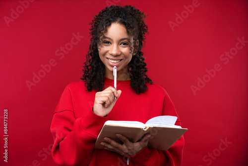Portrait of a young girl with notebook smiling at the camera while standing against a red background. 