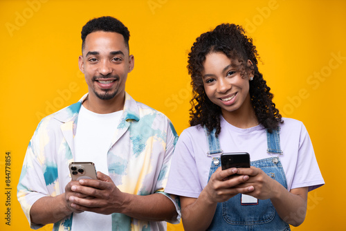 Two hipster friends smiling cheerfully while standing against a yellow background.