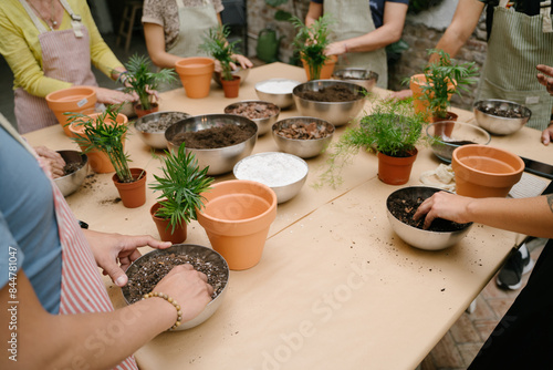 People at workshop class plant caring replanting house plants. table with soil, bowls, root balls, and plants. The hand is taking Perlite. Unrecognizable people hands