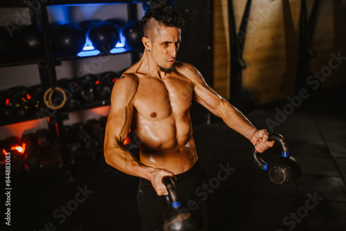 Shirtless handsome man covered in sweat working out with kettlebells in a gym photo