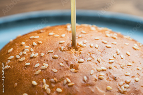 A wooden skewer stick is inserted through the center of the sesame seed bun, an extreme close-up shot. The bun is golden brown and appears freshly baked.