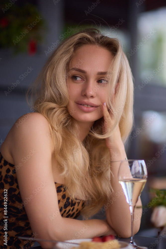A blonde woman in a leopard print top is sitting at an outdoor cafe, with a stylish city backdrop A blonde woman in a leopard print top is sitting at an outdoor cafe, with a stylish city backdrop